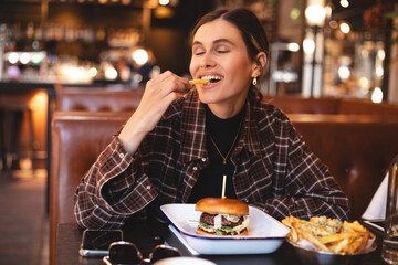 Fast food lover. Happy lady eating burger and french fries, biting potatoes and closed eyes, sitting in restaurant. Playful woman holding french fries, burger in plate and tasty. Cheat meals.