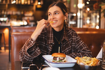 Fast food lover. Happy lady eating burger and french fries, biting potatoes and looking aside,...
