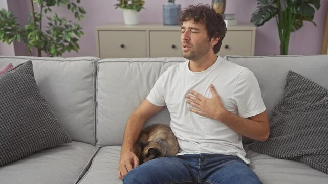 A young hispanic man feeling chest pain while sitting on a couch indoors with a siamese cat