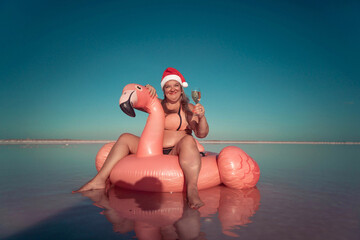 Santa Flamingo Christmas woman Riding Inflatable Flamingo Pink Lake. A woman wearing a Santa hat sits on a pink flamingo float in a body of water. He is smiling. © svetograph