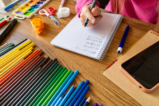 Child doing math test in notebook using smartphone with stationery on wooden table. Girl hand writing mathematical exercises as school homework. Learning, education using digital tools concept
