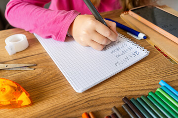 Child doing math exercises in notebook using smartphone with stationery on wooden table. Girl hand writing mathematical equations as school homework. Learning, education using digital tools concept.