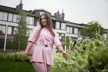 A young woman in a pink shirt and pants walks past a row of lush green bushes in front of a modern building