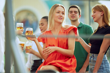 Group of friends enjoys lively outdoor gathering. Blonde woman in vibrant orange dress, holding drink and sharing laugh with her companions. Concept of Friday mood, celebration, Oktoberfest.