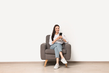 Smiling woman with smartphone sitting on armchair against white wall