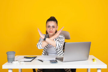 Showing stop gesture, young employee business woman sit office desk over yellow background holding...