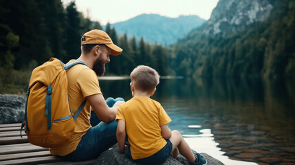 A bearded man and a young boy, both wearing yellow outfits, sit by a tranquil lake surrounded by a forest and mountains. The man has a yellow backpack.