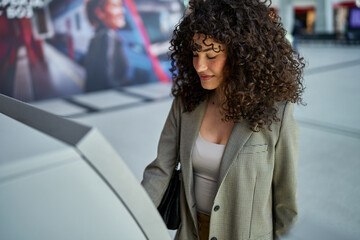 Elegant woman with a chic blazer using a street-side ATM, urban life in the background.