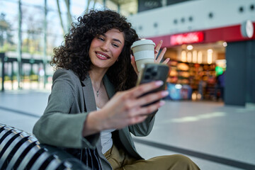 Smiling woman with curls taking a selfie at the train station, with a coffee to-go.