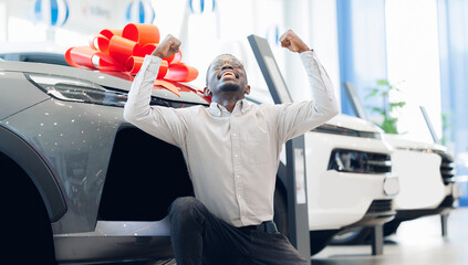 Handsome happy African American man is hug his new car and smiling in dealership