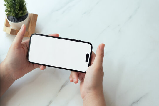 Hands holding a smartphone horizontally with empty screen over a marble table