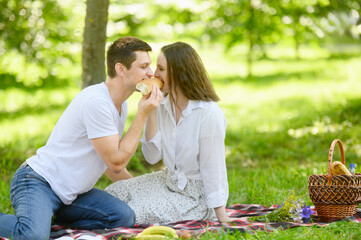 Man and woman enjoying delicious picnic in park, biting baguette together, surrounded by beauty of nature on sunny day. Romantic date of couple. Valentine's Day