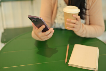 Cropped shot young woman with paper cup of coffee in hand using mobile phone in cafe