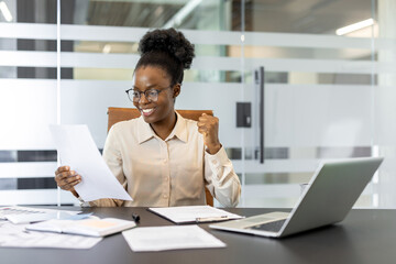 African American businesswoman rejoices at positive news while reviewing paperwork at desk. Bright...