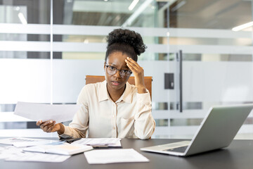 African American businesswoman appears stressed while working at desk with papers and laptop. She holds document and glasses, portraying workplace challenge and professional stress in office setting.