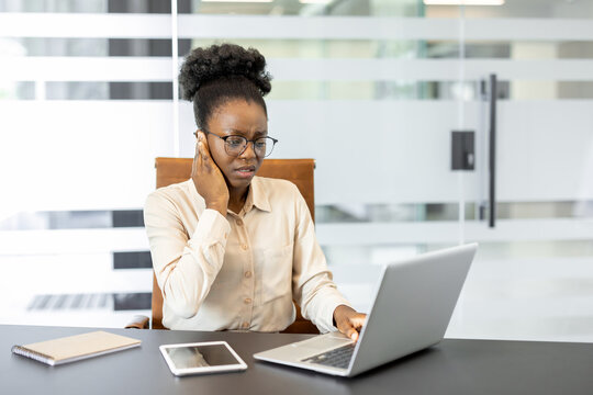 African American businesswoman experiencing ear pain due to inflammation, working in office. Woman uses laptop, probably facing discomfort. Includes phone and notepad