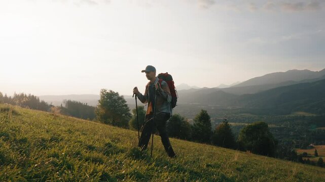Side view following man with red backpack and trekking poles walking on green grass meadow hill overlooking epic mountains and village in valley. Male backpacker explorer hiking, sunny summer morning