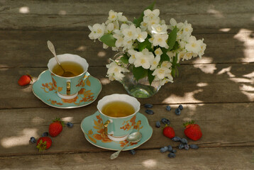 two turquoise cups of tea, a small bouquet of fragrant Jasmine flowers, with strawberries and honeysuckle scattered on the wooden table top