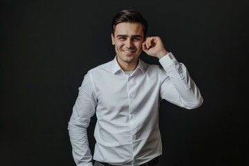 Fototapeta premium A confident young man in a white shirt stands against a dark background, offering a friendly smile while adjusting his ear thoughtfully.