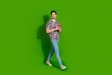 Full size photo of clever optimistic guy dressed print shirt jeans holding smartphone go empty space isolated on green color background