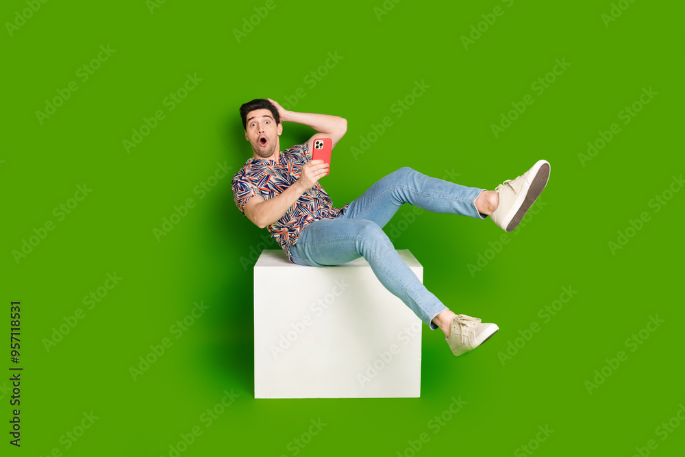 Wall mural full size photo of astonished guy dressed print shirt sit on white cube holding smartphone arm on he