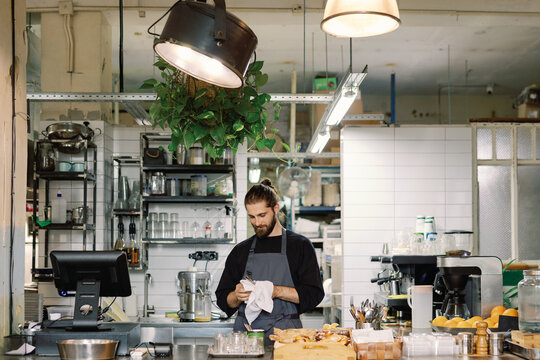 Bearded male cashier or waiter cleaning forceps with towel in cafe