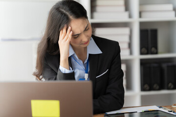 Overwhelmed Businesswoman: A woman in a suit sits at her desk, hand on her forehead, looking...