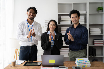 Business Team Applauding Success: A diverse team of three business professionals stand in a modern office, clapping in unison, their faces radiating joy and accomplishment. A laptop, paperwork, and co