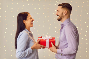 Happy couple in love standing together, holding red present gift box, looking at each other and laughing. Loving man and woman congratulating each other on holiday, sharing special romantic moment.