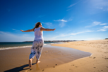 Beautiful middle aged blonde woman wearing white top and white pants with colorful patterns running barefoot with arms raised on sandy beach on ocean shore on sunny day. Happy holidays. Back view.