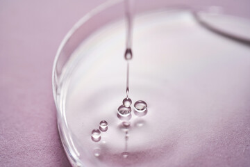 Serum or cosmetic oil flows into a transparent bowl on a purple background.