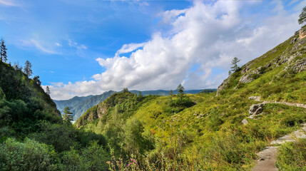 Fototapeta premium Panorama of a sunny summer landscape in the Altai Mountains. A narrow winding path leads upward. A cap of white clouds lies on the peaks of the mountain range in the background