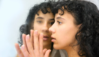 Sexy, young, seductive woman gently touches a mirror, her reflection staring back with deep...