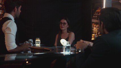 Focusing on beautiful woman guest sitting in special drink gin fizz at luxurious counter bar at vibrant nightlife party atmosphere. Smart bartender wiping glass while talking with customer. Vinosity.