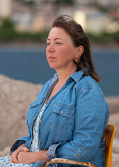 Portrait of  50 year old happy woman sitting on  chair.  Rocky seashore. Enjoying  vacation at the sea.  Travel and vacation concept