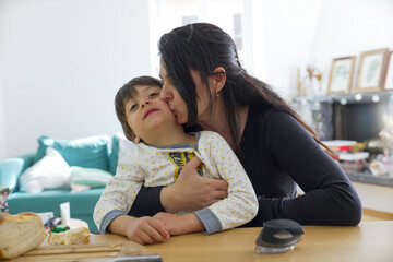 Mother lovingly kisses her son on the cheek while holding him at the kitchen table, creating a heartwarming scene. Bread and a kitchen utensil are visible, adding a homey touch.