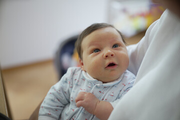 Newborn baby in light blue onesie with boats print, gazing up with curiosity while being held in arms, in a cozy home setting, highlighting early childhood wonder and family love