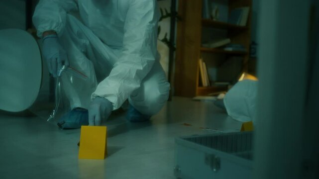 Low section of criminalist in coveralls disposable gloves and shoe covers crouching on floor and collecting bullets with tweezers into evidence bag at crime scene