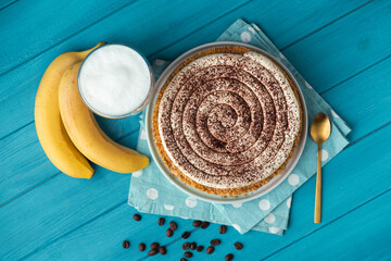 Top view of banoffe pie filled with banana and dulce de leche and sprinkled with grated chocolate. Banana cake on white plate with glass of coffee on turquoise summer background. Flat lay
