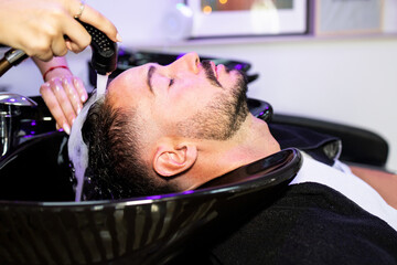 An unrecognizable stylist's hands wash the hair of a handsome young man with a beard in a sink of a modern hairdresser's. Hair washing concept before cutting.