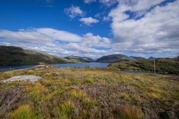 The image shows a grassy area with a body of water in the background. It is an outdoor landscape in Scotland, featuring a lake or loch, grass, and mountains in the distance.