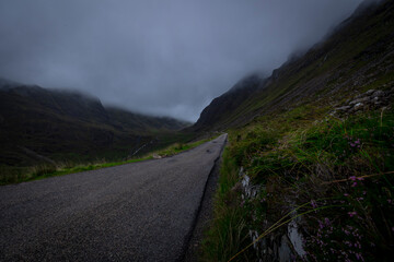The image depicts a scenic road winding through misty mountains, with clouds hanging low in the sky. The landscape features lush greenery and the silhouette of hills in the background. scotland