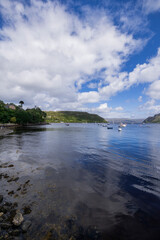 A serene landscape featuring a body of water with boats and buildings along the shore. The image captures a peaceful scene with a cloudy sky overhead.  Portree, Scotland, isle of skye