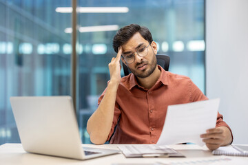 Stressed businessman reviewing documents at office desk during workday, experiencing confusion and frustration