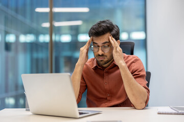 Stressed office worker at desk holding head in hands while looking at laptop screen in modern...