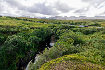 A stunning waterfall cascading down rocks in a lush green forest setting. The image captures the beauty of nature with a cloudy sky above. 
