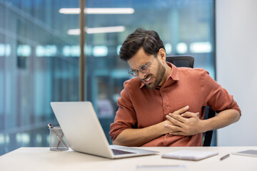 Businessman experiences chest pain while working at desk in modern office setting, indicating health issues during work hours