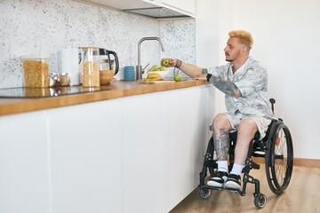 Caucasian man with tattoos sitting in wheelchair in modern kitchen preparing healthy breakfast with fresh green apples and other ingredients around him