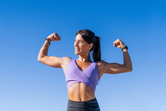 A young woman smiling and flexing her arm muscles under a blue sky