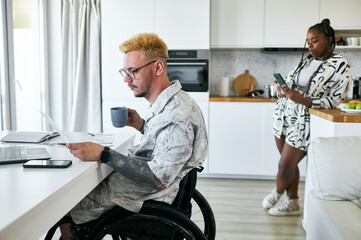 Man in wheelchair reviewing documents while holding mug, with woman standing in casual attire using smartphone in background, creating modern yet homey ambiance